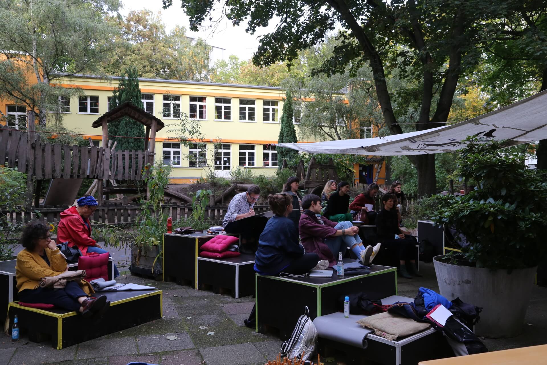 Personen sitzen in einem Außenbereich und hören einer sprechenden Person zu. Es handelt sich um Teilnehmer*innen des Netzwerks Access Point Tanz.